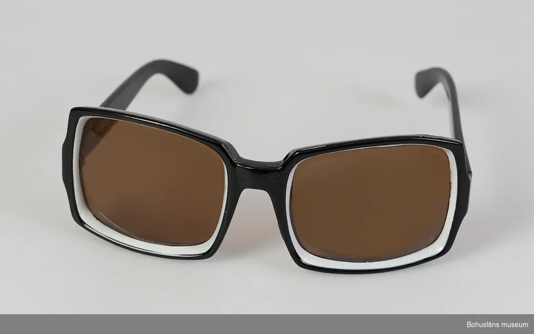 Ett par solglasögon, svarta med vit kant runt det bruntonade glaset. Både "glas" och bågar tillverkade i plast.
Köpta år 1985 i allt-i-allo-bod i Årjäng, Värmland av givaren.