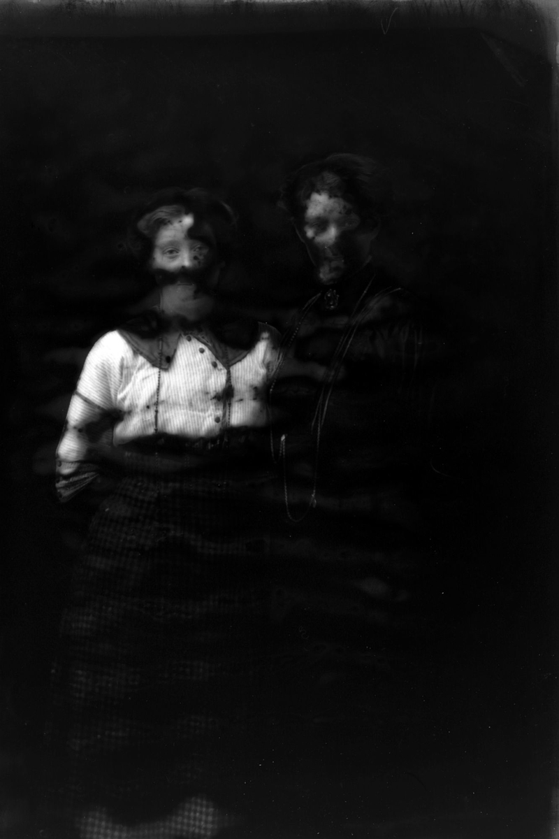 Studioportrett av to personer, begge trolig kvinner. Glassplaten til bildet er ødelagt.