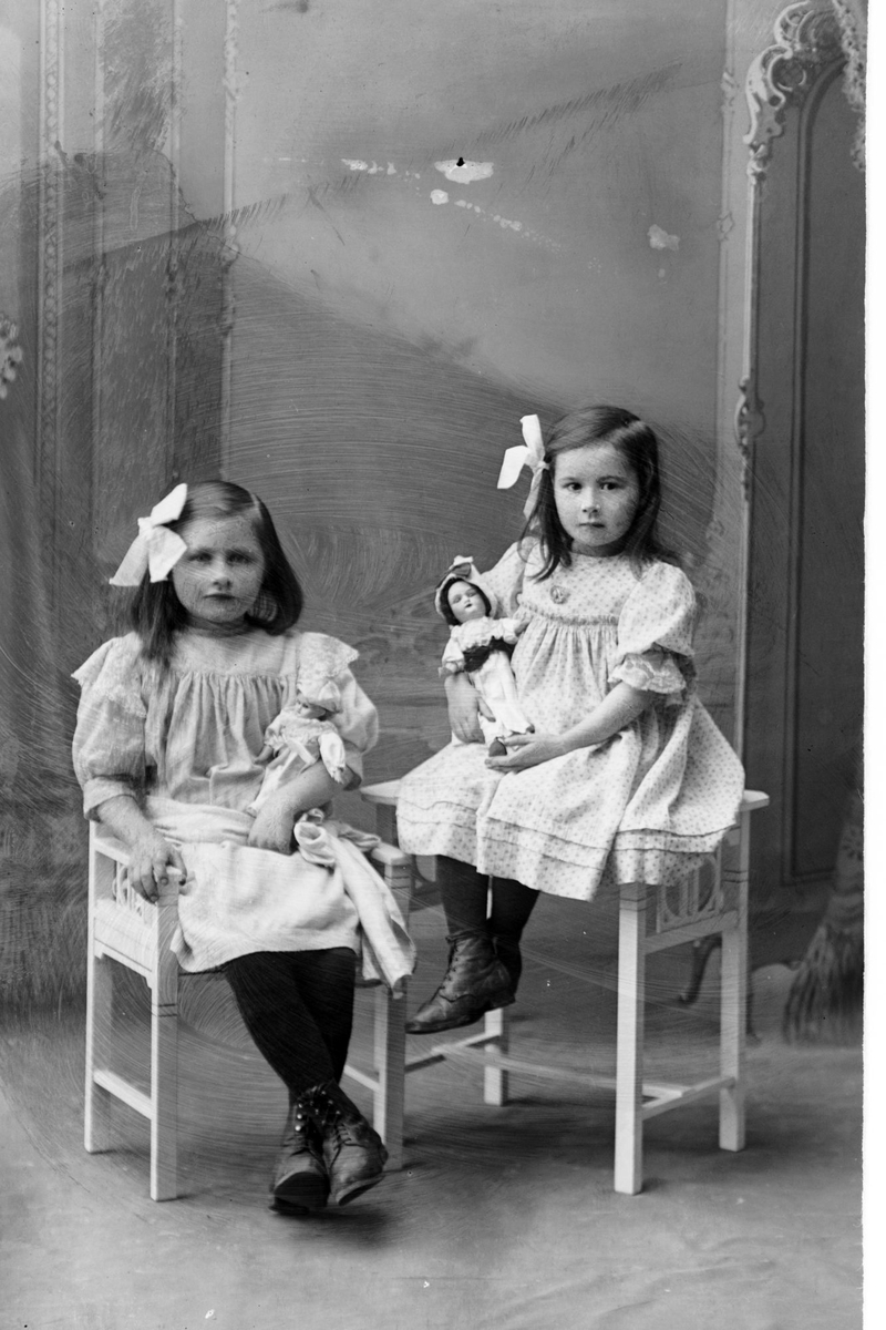 Studioportrett av to jenter, én med dukke i armene.