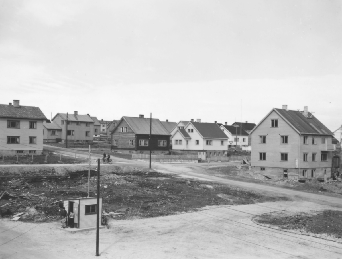 Motiv i fra gjenreisningen av midtbyen i Vadsø etter andre verdenskrig. På bildet ser man flere boliger. En tomt er tom. I den lille bua hvor det står noen personer utenfor var det drosjestasjon. Det er to syklister på grusveien ovenfor tomta