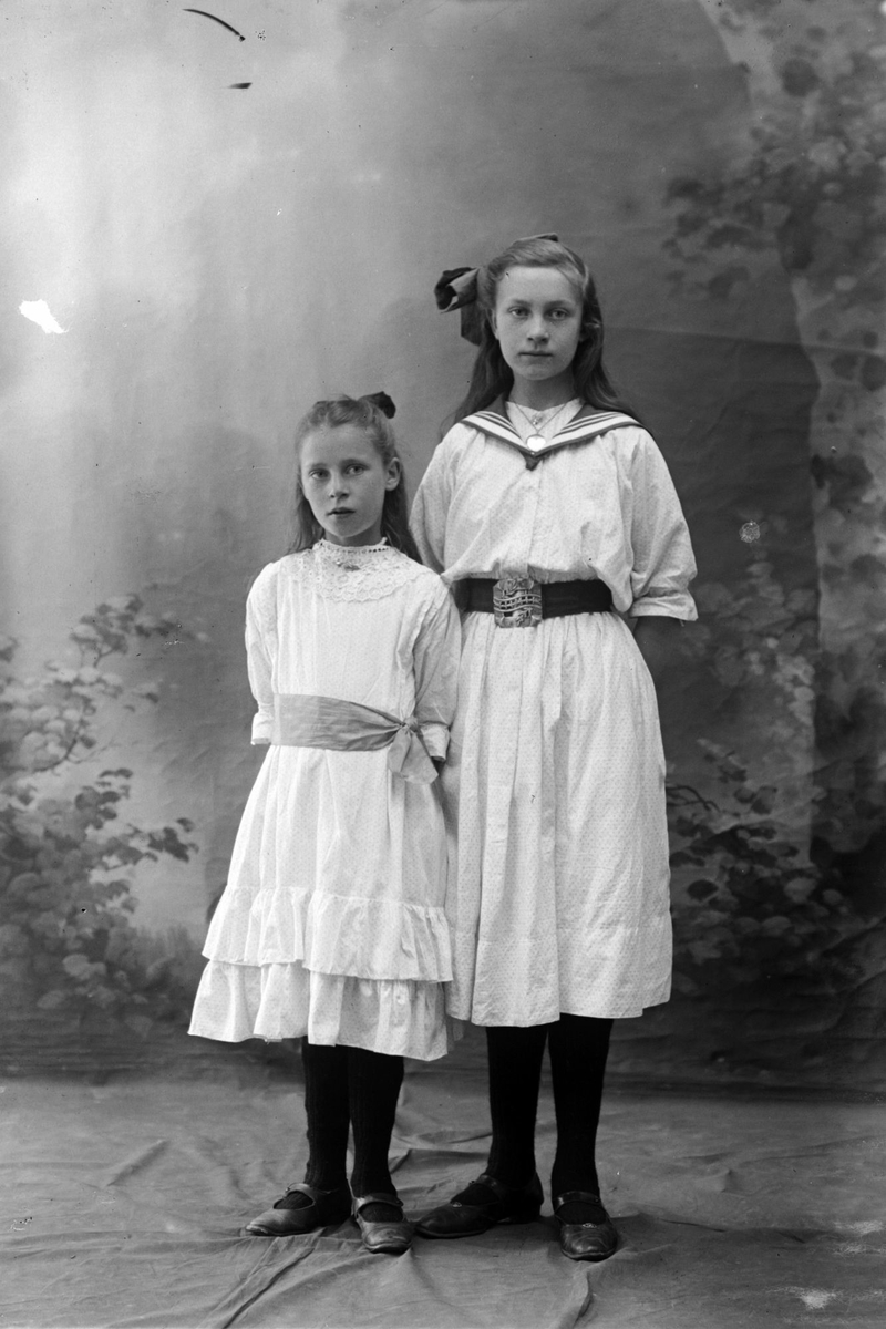 Studioportrett av to jenter som er kledd i lyse kjoler.