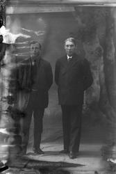 Studioportrett i helfigur av to menn. Bildet er delvis ødela