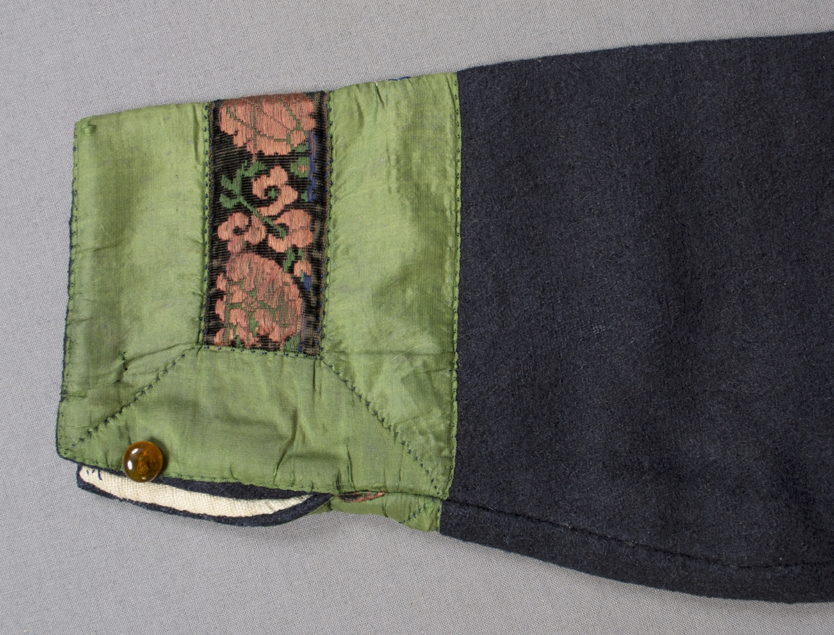 Blå vadmalströja garnerad med band och broderier. Tröjan har en rund öppning fram på bröstet och skört bak. Runt halsringning och öppningen fram, längs nederkanten på framstycken och ärmar är tröjan dekorerad med ett grönt 38 mm brett sidenband. Nederkanten på ärmen är dessutom dekorerad med ett mönstrat band i rosa och grönt silke på svart sammetsbotten. Framstyckena är också prydda med broderier på och intill det gröna bandet. Broderat blommönster finns också ovanför bandet på ärmen samt ovanför skörtet mitt bak. Framtill knäpps tröjan på två ställen med en hyska och hake. Ärmlängd: 480 mm. Ärmen är sydd i ett stycke samt en kil. Ärmsprundet är 80 mm långt och knäpps med en rund bärnstensknapp, tränsat knapphål. Axelbredd: 120 mm. Bakstycket har söm mitt bak. Tröjan är helfodrad med handvävt, tuskaftat halvblekt linne. Broderierna är utförda i plattsöm och flätsöm i olika nyanser rosa och gult samt i blått, vitt och grönt silke. Foder och band är påsydda för hand.