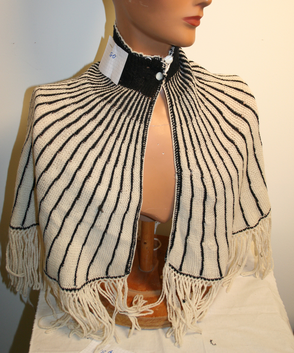 Skuldersjal, sengehygge i ull, strikket i hvitt med svarte striper, med hvit heklekant og 12 cm frynser nederst. Knepning i halsen.