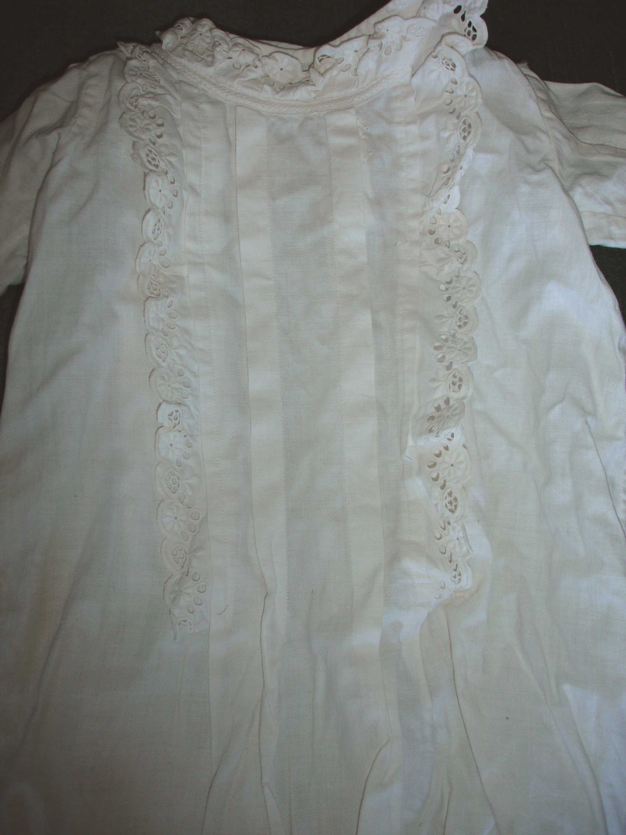 Dukkeklær. Kjole i hvit bomull med blondekant som kappe rundt halsen og langs kanten foran, samt nederst på ermene. 