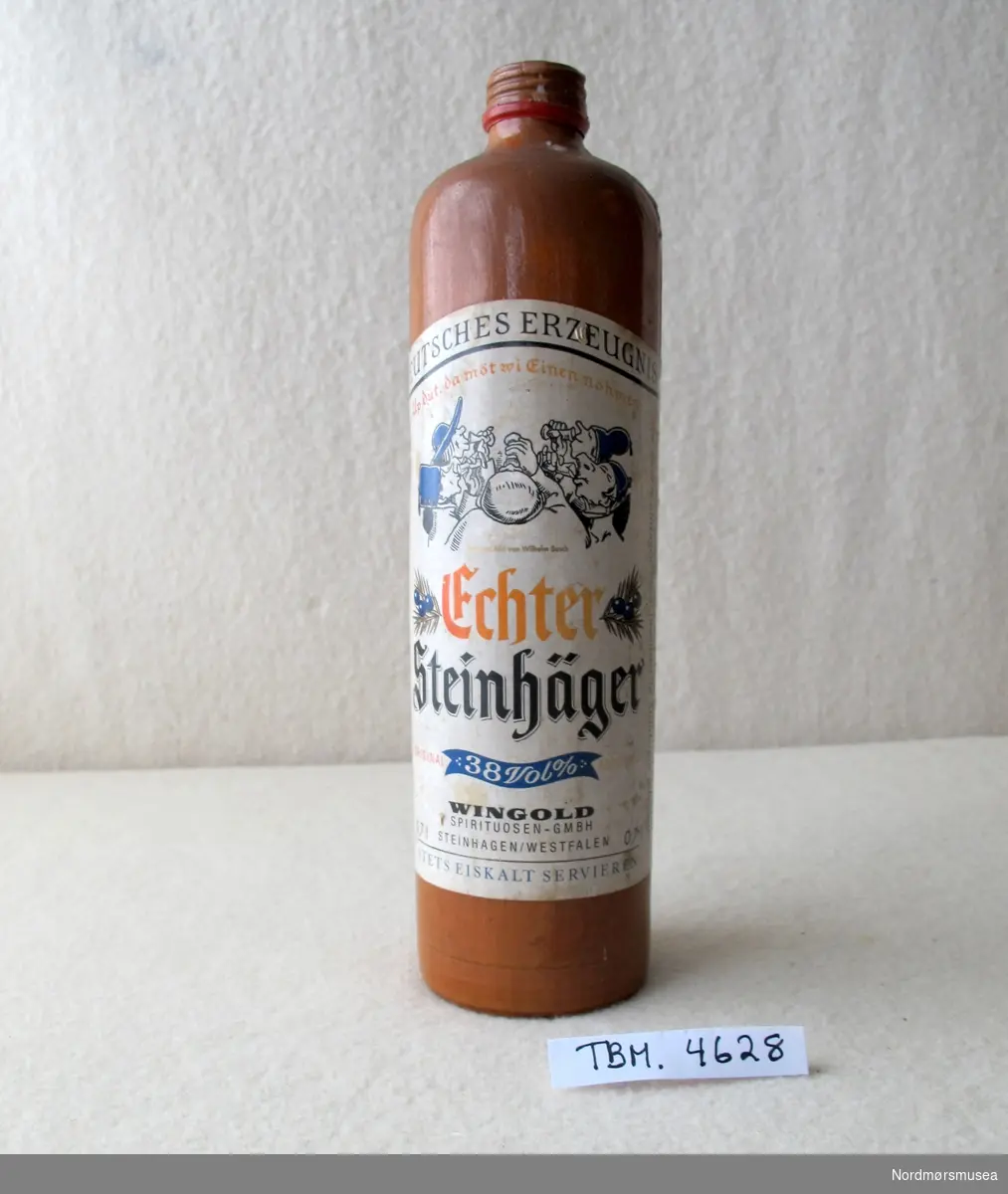 Røyrforma flaske i brunt leirgods med etikett av papir. Tekst og bilde på etiketten.