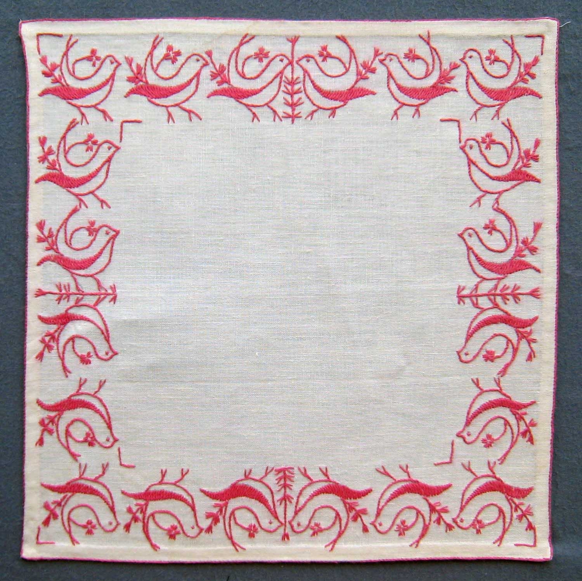 Fyrkantig duk i vitt linne broderad i Melpasöm med rosa dubbelt garn, 1 tråd lingarn och 1 tråd bomullsgarn.
Fåglar i stjälkstygn, flätsöm och tofssöm runt kanten.