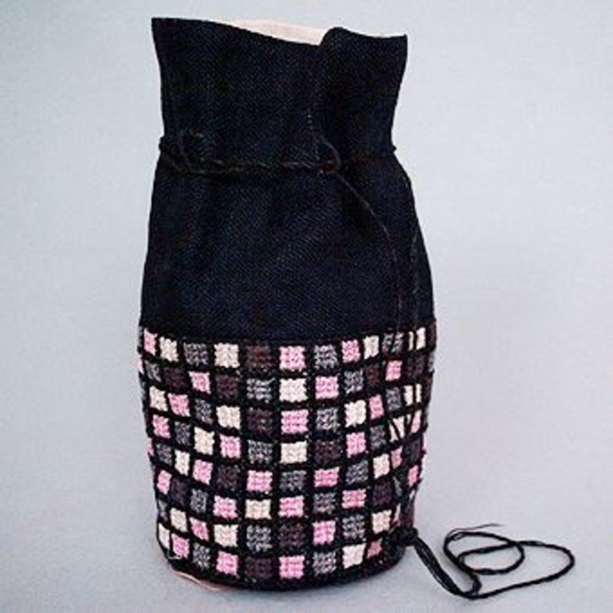 Liten väska med nederdelen broderad i rutmönster med rosa, grå, bruna och halvblekta rutor i tvistsöm och överdelen i svart linnetyg.På etikett fastsatt i botten på väskan anges materialåtgång och prisuppgift, texten är svårläslig."Skollinne nr 1 35 x 15 cmSvart linne 30 x 15 cmVitt linne t foder 35 x 25 cm"Knox garn och Hålsömsgarn har använts"Knocks 1 docka rosa 556Knocks 1 docka grå 524Knocks 3 dockor svart 5911 1/2 docka grön?hålsömsg. nr 5?Pris 10 kr"."Modell får ej säljas".Väskan har SHR:s pappersetikett från Kronobergs Läns Hemslöjdsförening Växjö, med påskrivet nummer: "1409", fästad vid sig.