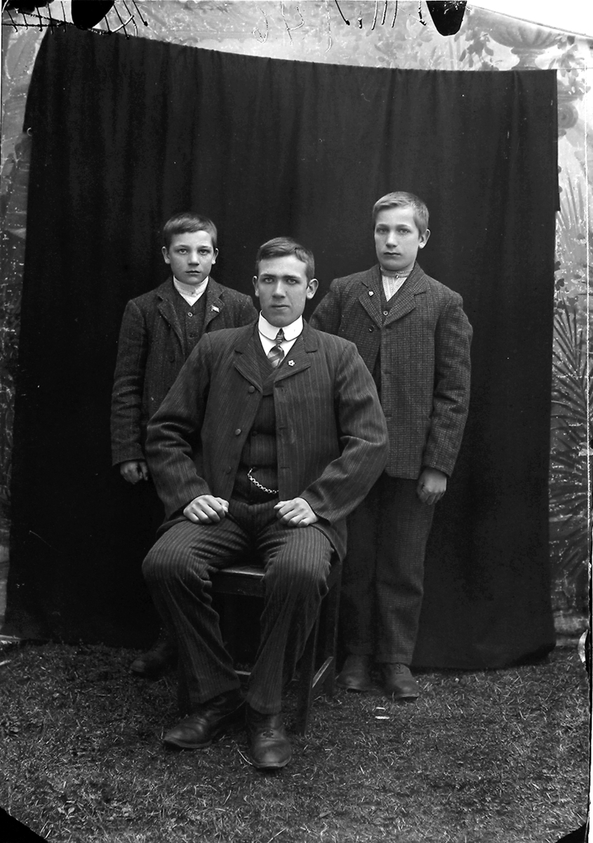 Portrett. 3 personer, alle i dress. En mann i 20-åra sitter. Bak ham står to gutter ca. 12-15 år. Bildet er tatt på Ramberg i Lofoten.