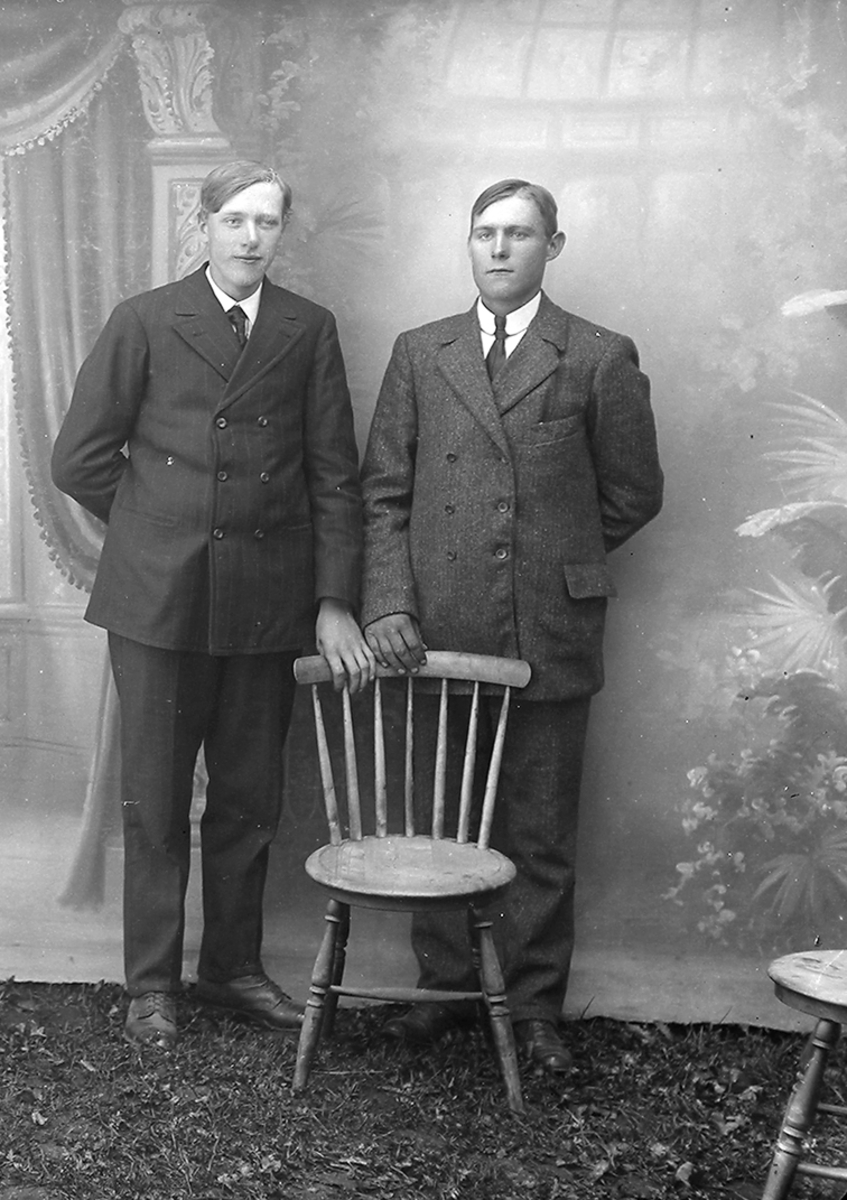Portrett. To menn. Står ved en stol. Begge har dress på seg.