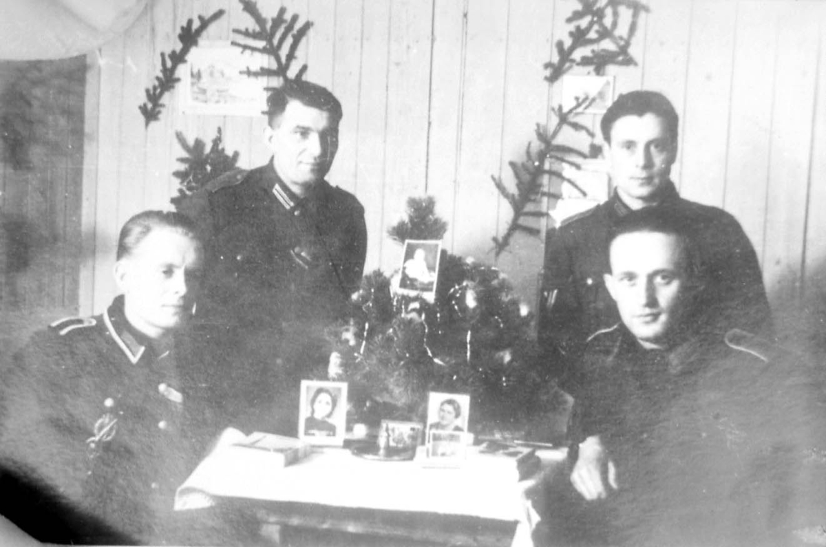 Gruppebilde. 4 personer, menn i militæruniform ved et bord. Noen fotografier på bordet. Innendøes.