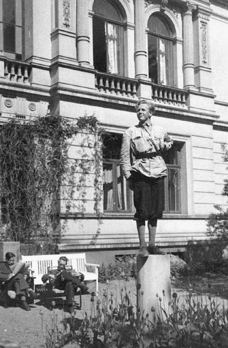 En person. Mann i uniform. Står oppå en sokkel. To personer sitter i bakgrunnen foran en bygning.