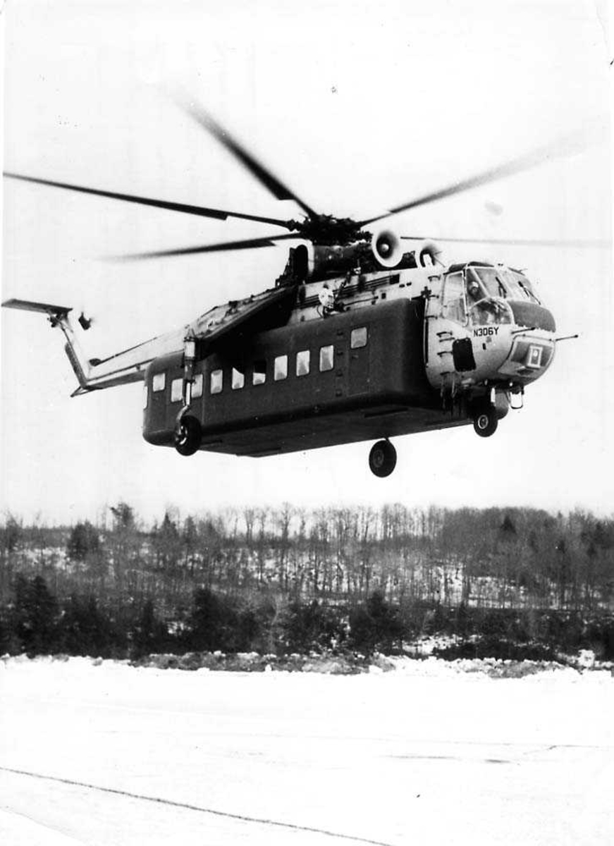 Luftfoto. Ett helikopter i luften like over bakken, Sikorsky S-64 Skycrane med en passasjerkonteiner festet til helikoptret, N306Y.