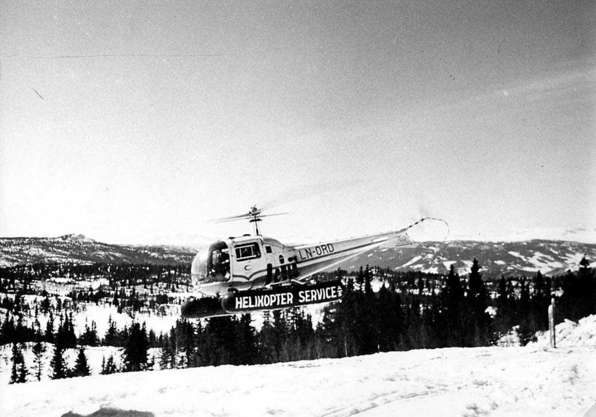 Luftfoto. Ett helikopter i luften, Bell 47 J Ranger LN-ORD fra Helikopter Service A/S. Fjell i bakgrunnen. Snø på bakken.