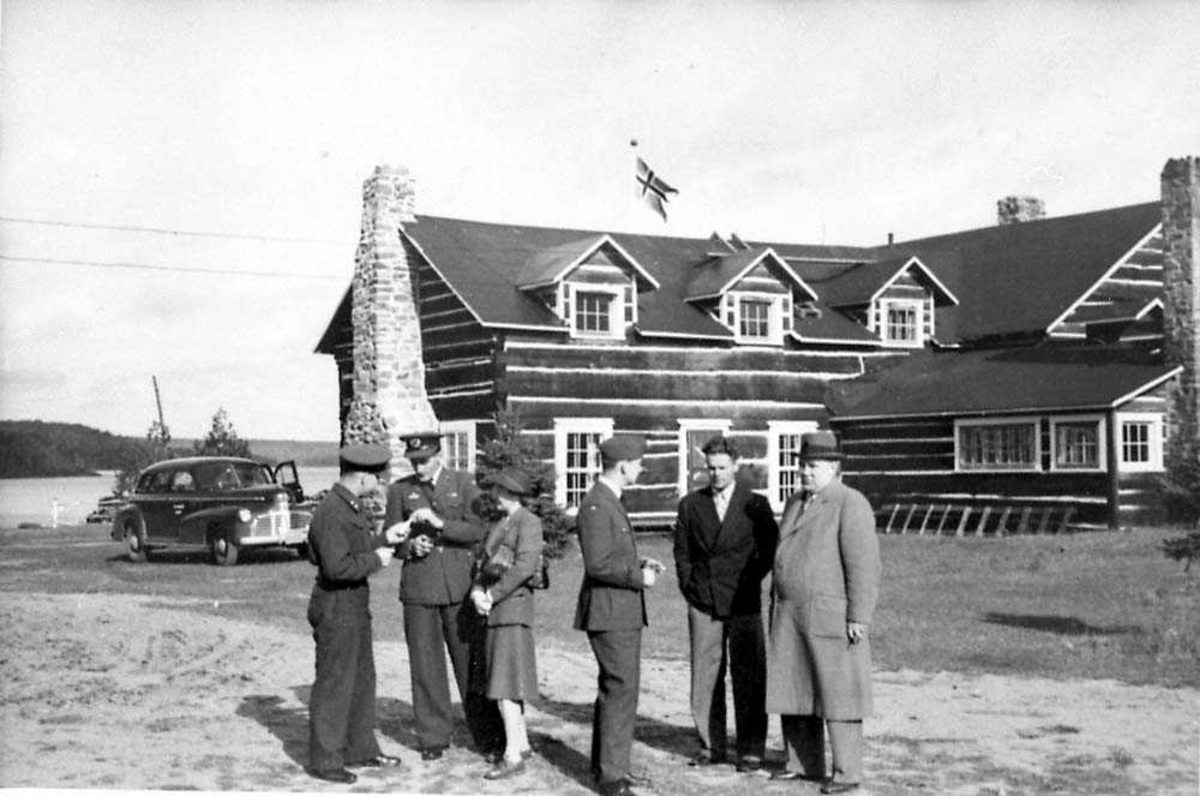 Seks personer som står på en åpen plass foran en bygning, fire av personene har militæruniform på seg.