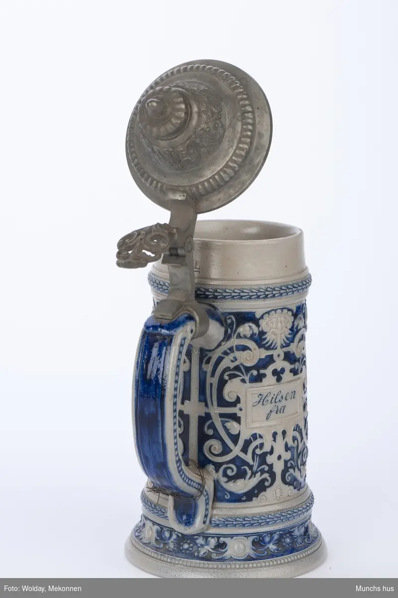 Drikkekrus av Hollands type. Blågrått stentøy med blå dekor. 
Barokkinspirert dekor.
Lokk i metall.