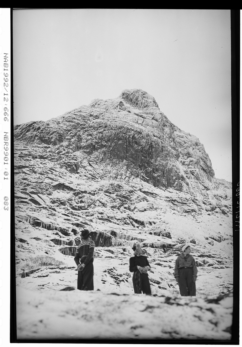To menn og en kvinne står ved fotenden av snødekt fjellside. Ukjent.