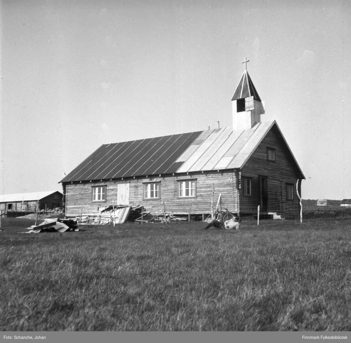 Et bilde fra kirken i Kautokeino. En mann ligger på gresset og koser seg i sola foran kirken. Ved siden av ligger byggematerialer, taket på kirken er under reparasjon