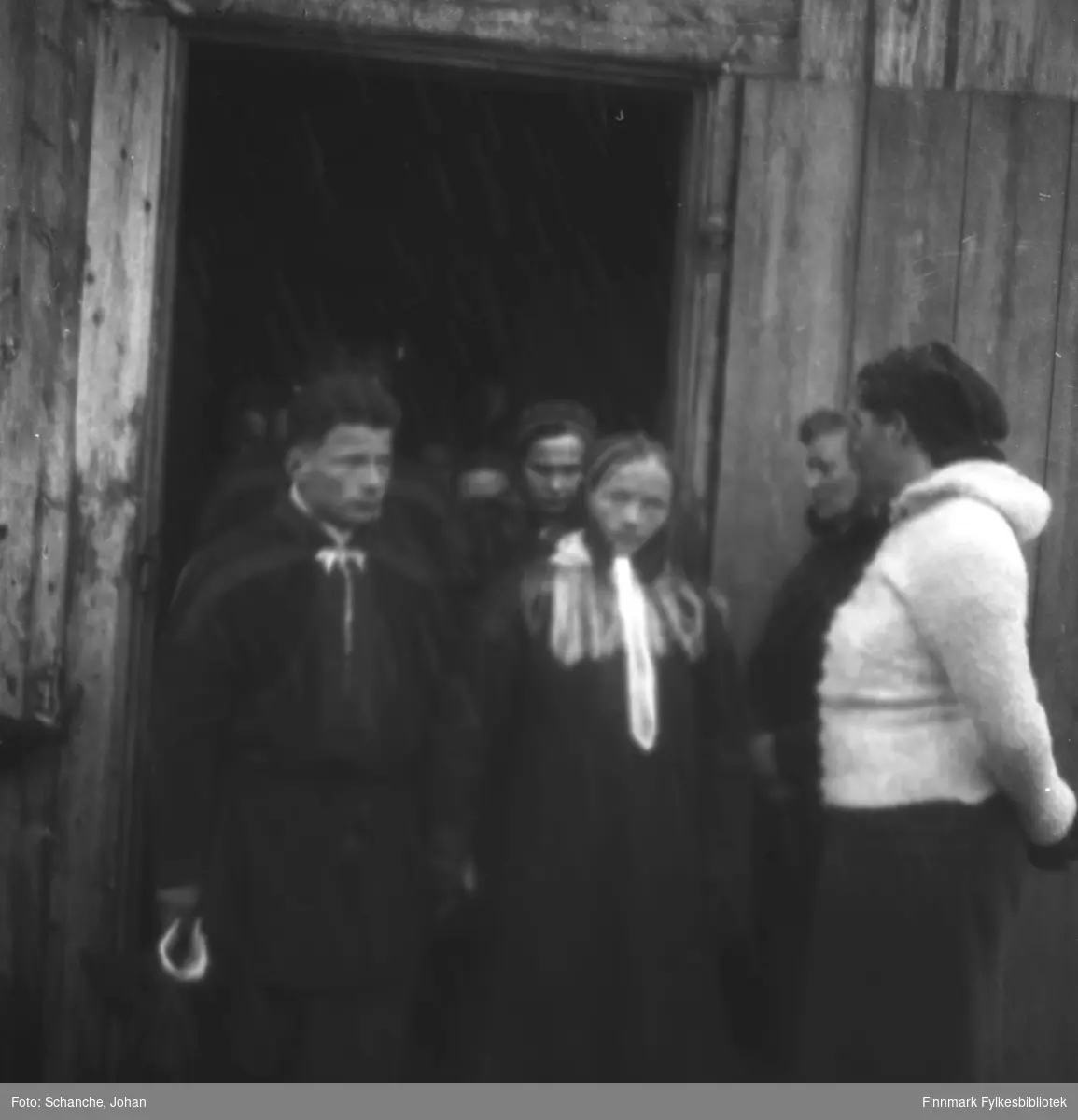 Menn og kvinner i samedrakt kommer ut av Polmak kirke påsken -46.  Bildet er tatt ved døråpningen, to kvinner står ved paret i sametrakt som komer ut av kirka. Muligens brudepar?