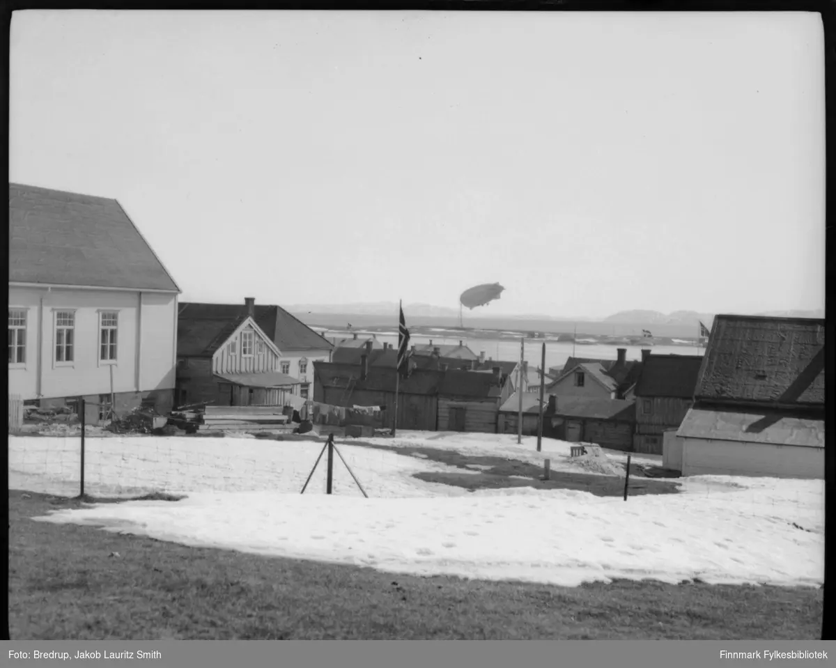 Dette er antakelig luftskipet Norge forankret ved luftskipsmasta på Vadsøya.  Bildet er tatt mellom bygninger i Vadsø fra samme vinkel som FBib.98012-016, som vi gjetter er tatt under Italias besøk i 1928.  Det ligger mye snø og man flagger i byen