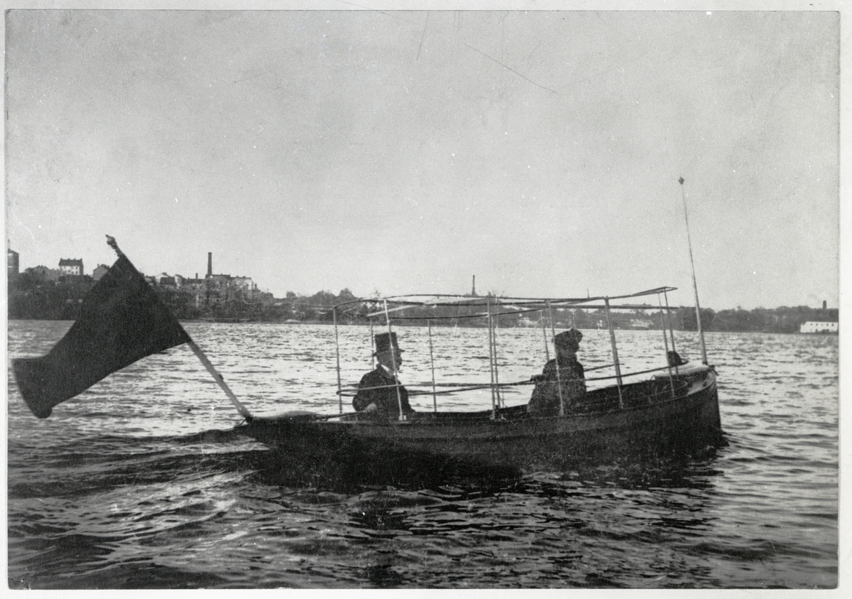 [från fotobeskrivningen:] "Mannen till rors i båten iförd hög hatt är Henning Forslund." "Båtens längd 7 m Maskin 1 cyl. 3 hkr motor Fart 5,5 knop" "Föreningen Sveriges Sjöfartsmuseum i Stockholm No. A 988"