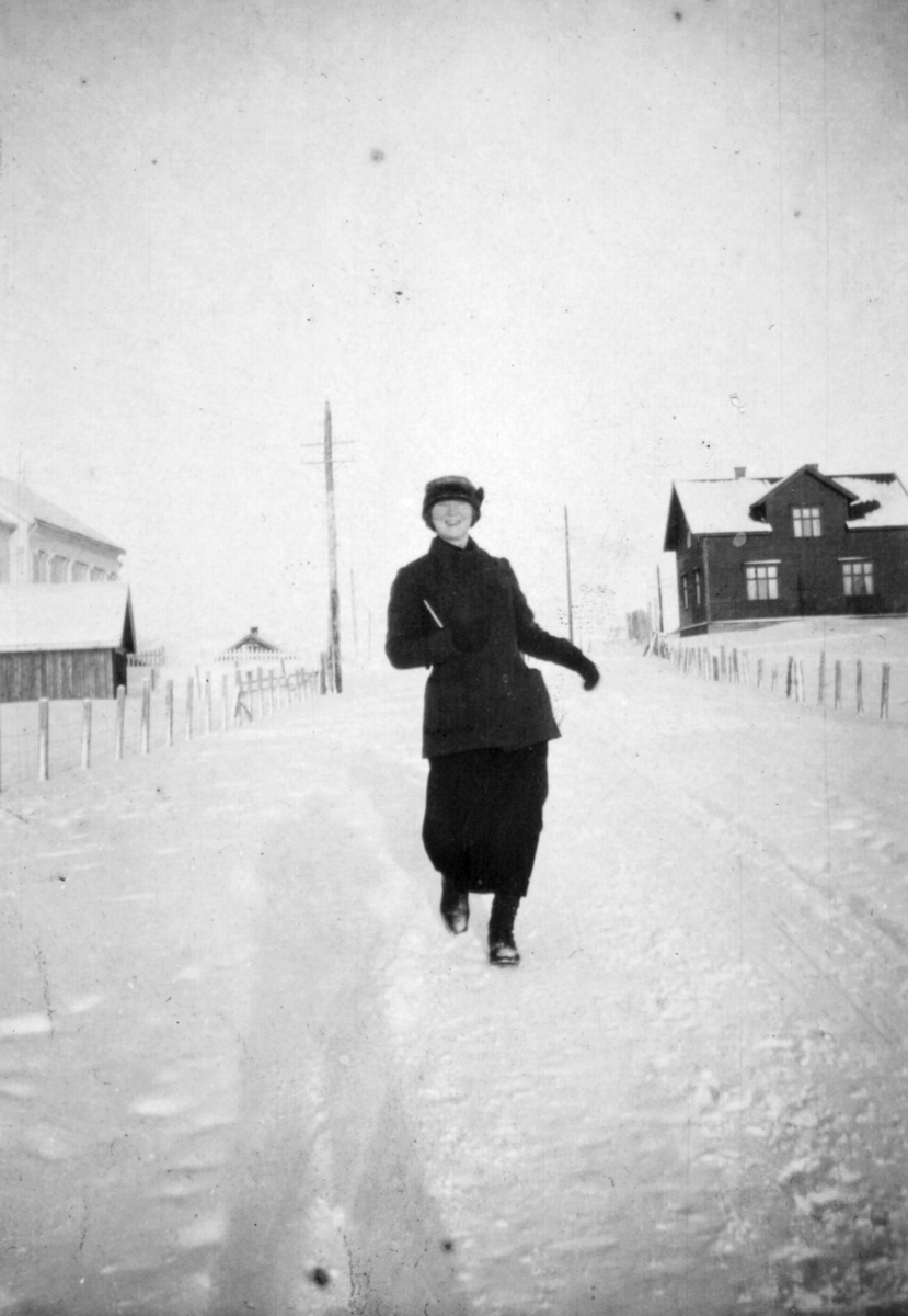 Sara Gabrielsen fjoller seg og går i snøen mellom Tana kirke til venstre og klokkergården til høyre, Langnes i 1916.