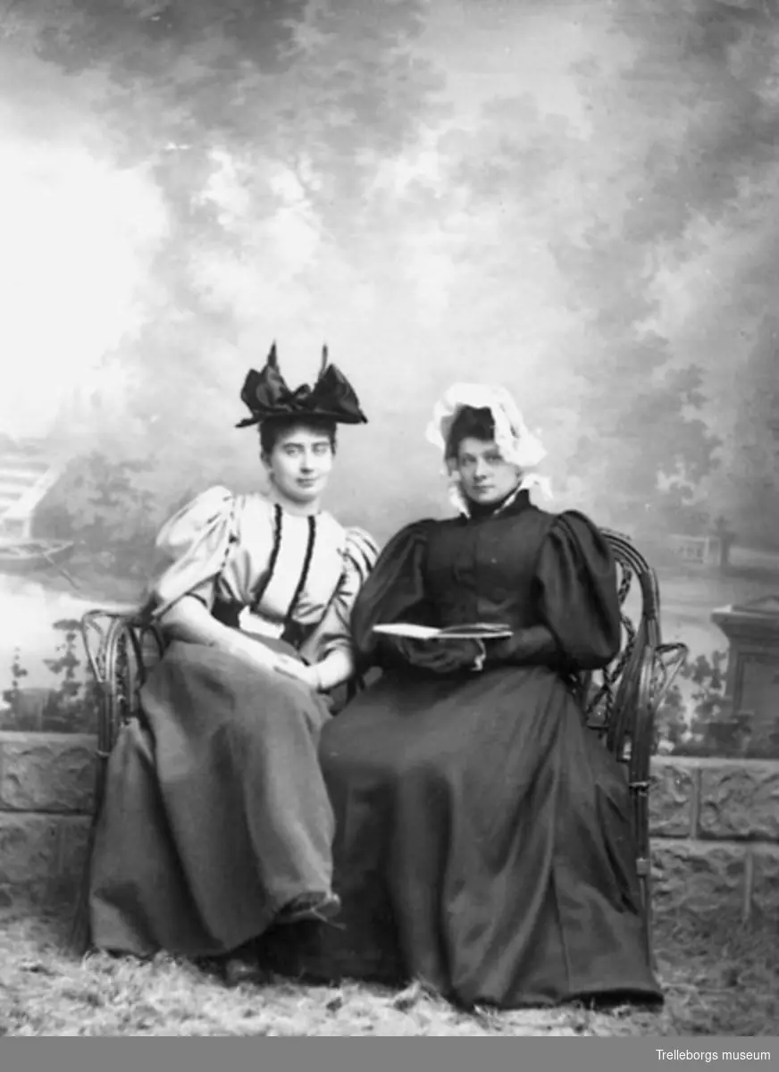 Två kvinnor, en i svart klänning med vida ärmar och vit spetsmössa och den andra i hatt prydd med en stor rosett.