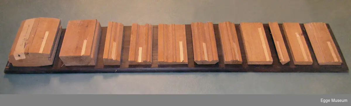 Rektangulær granplank påmontert ubehandlet og høvlet listverk og panel i furu og gran. Granplank er beiset brun og har sider/kanter som er avrundet. De påmonterte bordene er skrudd fast til beiset granplank. Alle bordene er produsert ved Bangdalsbruket (nedlagt i 1980).

1. Plank i gran, til maskinlaft av hus, pløyd med buet front, 195mm x 70mm. 
2. Fotlist i furu, grov, ant. for golv i arbeidsrom, 145mm x 47mm. 
3. Taklist i furu, profilert, grov, 65mm x 35mm.
4. Kombinasjonslist i furu, for dør/vindu, 90mm x 20mm. 
5. Dørlist i furu, profilert, 115mm x 15mm.
6. Taklist i furu, profilert (barokkaktig), 94mm x 20mm.
7. Dekorasjonslist i furu (karmlist?), profilert, 65mm x 23mm.
8. Glattkantet bord i gran (planhøvlet), 92mm x 17mm.
9. Hjørnelist i furu, grov, 40mm x 22mm.
10. Dekorasjonslist i furu (karmlist?), profilert (kan også være veggpanel uten not og fjær) 67mm x 17mm.
11. Panelbord i furu, for vegger og tak, profilert, 90mm x 17mm.

Brukt i undervisningen ved Skogskolen på Steinkjer.