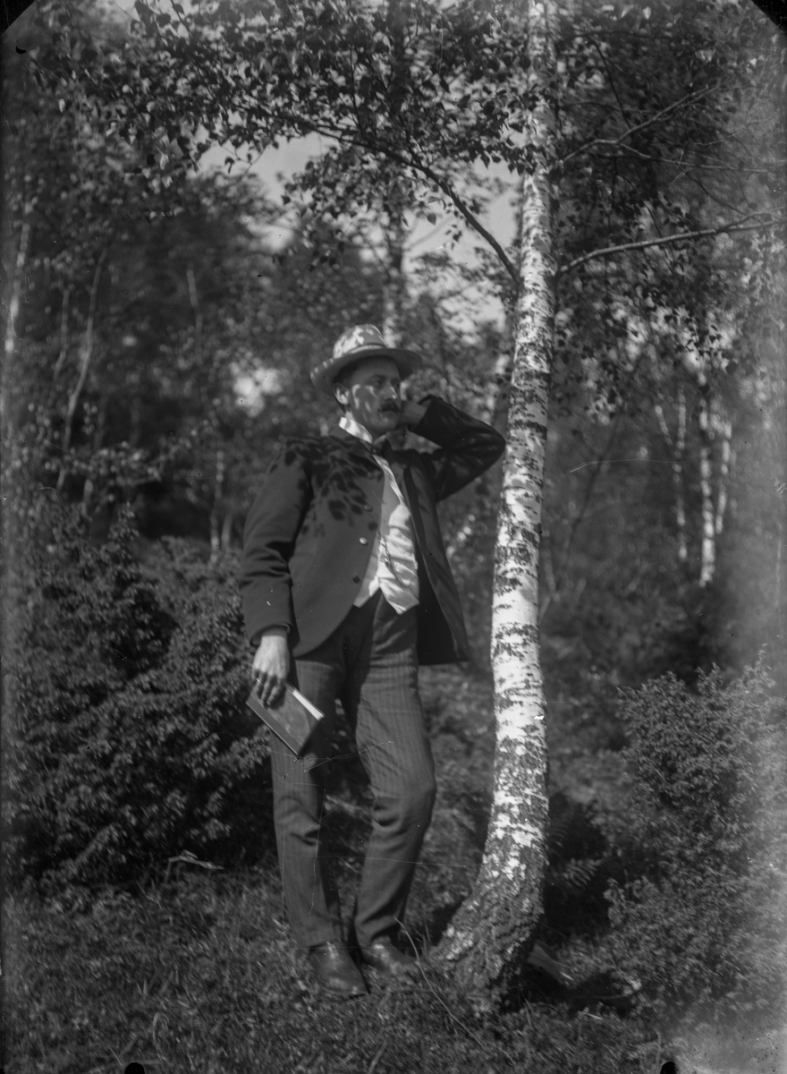 En mann med hatt ved siden av et tre, med flere trær rundt seg og i bakgrunnen. I sin høyre hånd holder han en bok. En busk til høyre.