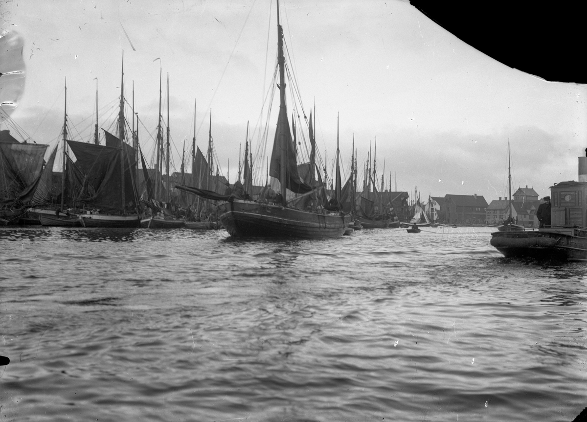 Mange sildefartøyer i Smedasundet. En færing omtrent midt på bildet. Et annet fiskeskip til høyre. Sjøhus i bakgrunnen.