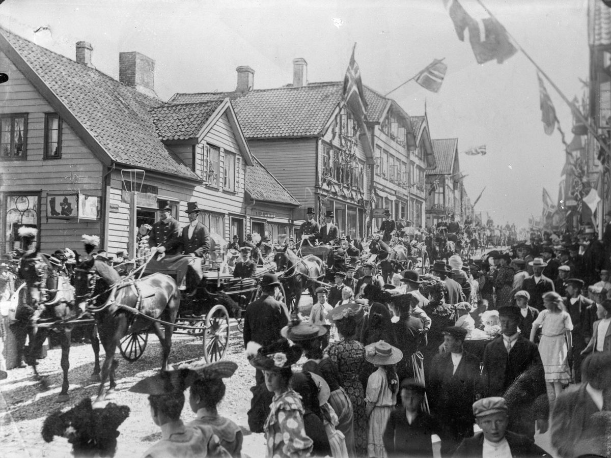 Fra kongebesøket 26. juli 1906. Haraldsgaten. Kongen og dronningen sitter i en vogenen som trekkes av to hester lengst til venstre. Bak dem følger flere hester med vogner hvor det sitter to personer på hver, i gaten. Mange personer på begge fortauene.  Trehus i bakgrunnen til venstre. Norske flagg.