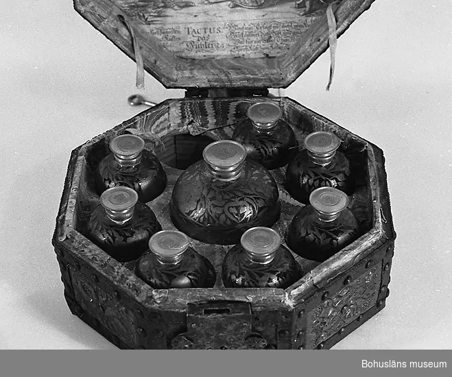 Ett åttkantigt skrin för nio glasflaskor. Skrinet är ett holländskt arbete från 1600-talet.
Skrinets utsida är beslagen med mässingsplåt, ca 0,2 mm tjock och hålls på plats med smidda band och prydnadsband av järn. Mellan banden framträder dekorativa rektangulära mässingsfält. Lockets två parallella band är klippta i ett löpande åttakantigt mönster passande skrinets form. Mässingsplåten är rikt dekorerad med blommor, hjärtan, cirklar och löpande linjer vilka formats med puns eller pressats fram.
Invändigt är skrinet rödbemålad i dess undre del med vad som troligen är tunn oljefärg, alternativt limfärg. Skrinets lock och den övre hälften av skrinets underdel är klistrat med papper av två slag. Locket pryds av ett kopparstick av amoröst slag som utgör en mindre del av en större bild. Texten "Tactus das fuhler", troligen motivets titel, är urklippt och placerad i anslutning till motivet. Ytterligare text beskriver kärlekens nöjen. Under titeln står "zu finden bey Joh. Peter Wolff in Nurnberg".
Övriga partier av ett marmorerat papper av holländskt slag. Marmoreringen är utförd med kam i rött, blått, vitt och gult. 
Lock och underrede hålls samman av två smidda gångjärn. I uppfällt läge har locket hållits på plats med två rosafärgade linneband.
Skrinets lås har låsbricka av järn dekorerat med ciselerade bladslingor.
Skrinet har kunnat bäras i ett handtag av smidesjärn, fästat ovanpå skrinets lock.
Inuti skrinet står sju mindre flaskor av blåst glas, (höjd 11 cm, diameter 7 cm) samt en större flaska (höjd 13 cm, diameter 10 cm). Flaskorna har svarvade och stämplade skruvlock av tenn. Flaskornas skuldror är dekorerade med bladguld lagt i olja och föreställer blad, blommor och torligen frukter.
Utförande är ganska grovt med obehandlade geringar, och sammanfogning med mindre spik.
Om Johann Peter Wolff finns uppgifter att han gjorde kopparstick på 1700-talets början.
Ur handskrivna katalogen 1957-1958:
8-kantigt skrin med 8 glasflaskor [en flaska saknas av de nio platser för flaskor som finns]. Skrinets L. 30,3 Br. 30,3 H. 16,5. Utvändigt klätt med järn- och kopparbleck [ska vara mässingsbleck]. Invändigt ställ för flaskor. Skrinet något skadat. Enstaka maskhål. En större flaska diam: 9,5. H.: 13. 
7 mindre flaskor. 5 st.: diam. 6,2. 2 st.: diam.: 6,0. Samtliga H:11. Med guldmönster och hattar av vitmetall. Hela.

Lappkatalog: 61
Ur handskriven katalog över Uddevalla - Musei Historiska - samlingar: Utländska föremål = De etnografiska föremålen upprättad  år 1916 av Knut Adrian Andersson, Intendent från 1 Oktober 1915:
No 22. Rese-Skrin, åttasidigt; utvändigt konstrikt beslaget med järn- och mässingsnaglar, inuti har det 8 glasflaskor med hattar av vit metall märkta L och däröver kunglig krona. Skänktes 1864 av Disponenten vid Kampenhofs fabrik i Uddevalla Robert Wilhelm Bley. (=G:23 i 1869 års tryckta katalog.)

Litteratur:
Framtidens Skrin, 300 års design, Hemslöjd Västra Götaland, s. 13 (Medicinskrin från 1600-talet; RMVG21609).
Wallin, Sigurd: Nordiska museets möbler från svenska herremanshem. Del I, 1500-1600-talen. Renässans och barock. Nordiska museet 1931 - 1935. Facsimile-upplaga 1979.