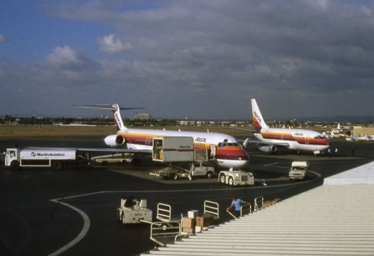 Lufthavn/Flyplass. To fly fra flyselskapet AIRCAL, USA, oppstilt ved flyplassterminalen. Stor aktivitet rundt flyene.