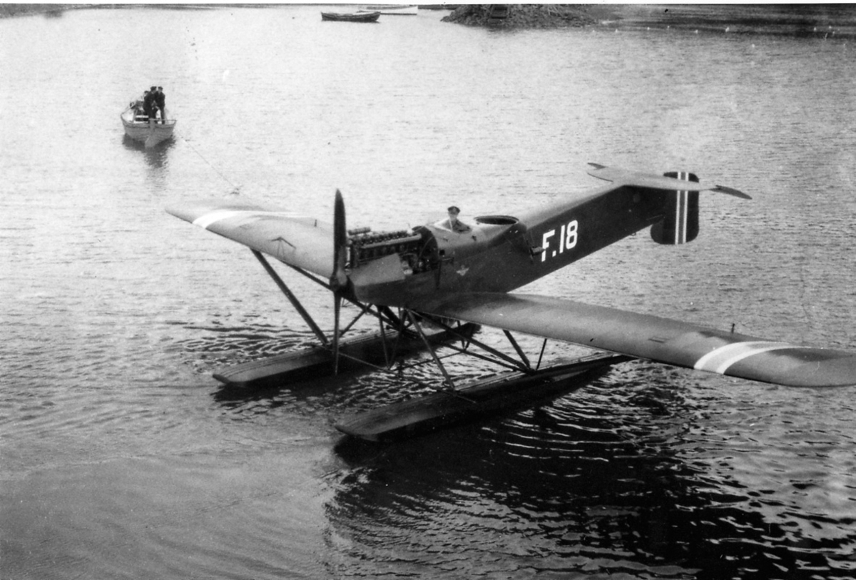 Ett fly, Hansa Brandenburger Nr. 64-F18.  en person i cockpit. Noen personer i en båt lenger bak.