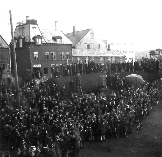 Frigjøringsdagene i Bodø etter krigen 1940 - 1945. Mange personer samlet nede på kaiområdet. Bygning bak med påskrift "CAFE". Kullkranen til Jakhelln t.h.