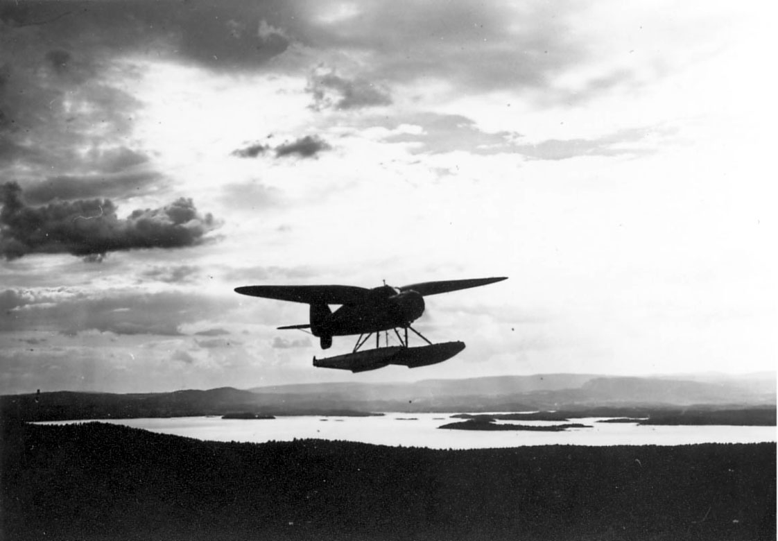 Luftfoto, ett fly i luften, landskapet sees nedenfor. Stinson SR.8EM fra Widerøe.