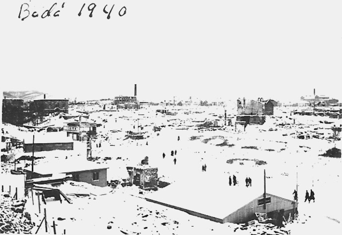 Bodø i ruiner etter bombingen under 2. verdenskrig. Snø på bakken. Flere personer blant ruinene. Noen brakkebygninger. Deler av torget, bryggeriet oppe til høyre.
