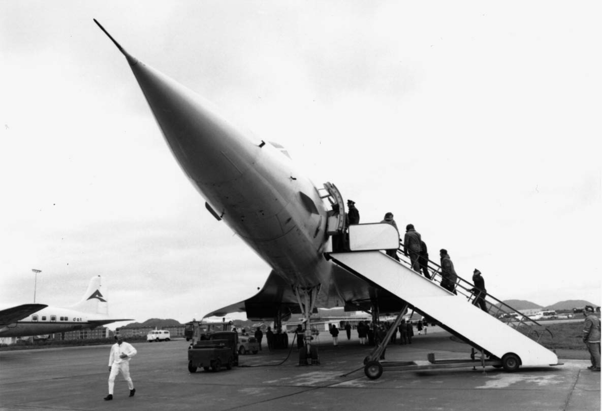 Lufthavn, 1 fly på bakken Concorde F-WTSB fra Air France. Noen persone/passasjerer på vei inn i flyet. Annet fly i bakgrunnen.