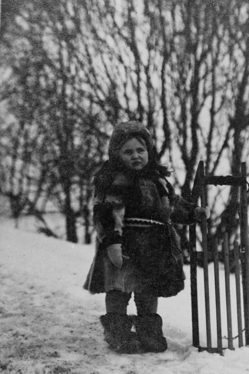 Lita jente i pelsklær står med en kjelke, fotografert i snøen utendørs.