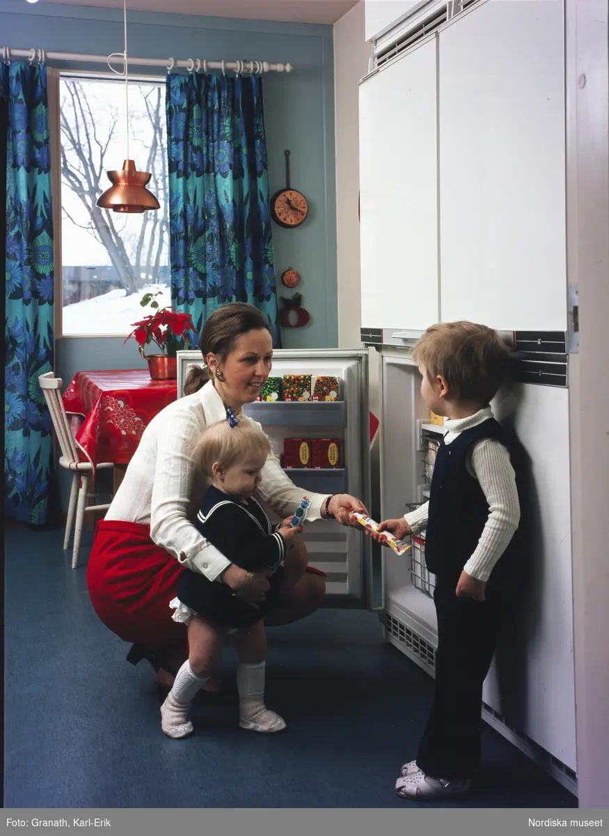 Modellbild i en köksinteriör. En kvinna sittande på huk vid ett kyklskåp med  två små barn- ett i sjömanskostym, ett klätt i  fluga och vit skjorta .