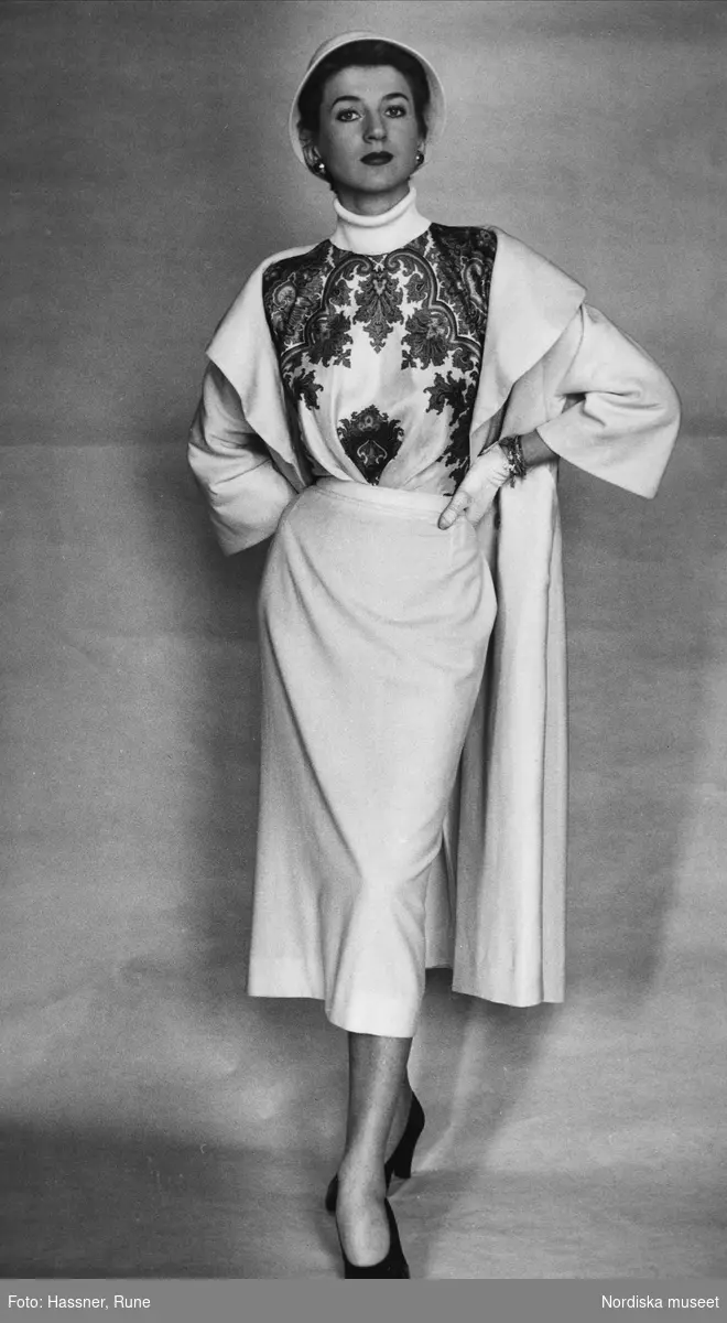 Modebild från Nordiska Kompaniet. "Italiana." Modell klädd i vit kappa, vit vadlång kjol, blus med polokrage, svarta pumps, vita handskar och vit hatt.
