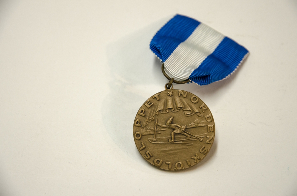 Idrottsmedalj i metall med motiv: Skidåkare. Medaljen har ett blå, vitt band och kommer från Nordenskiöldsloppet. På baksidan står ingraverat 1973.