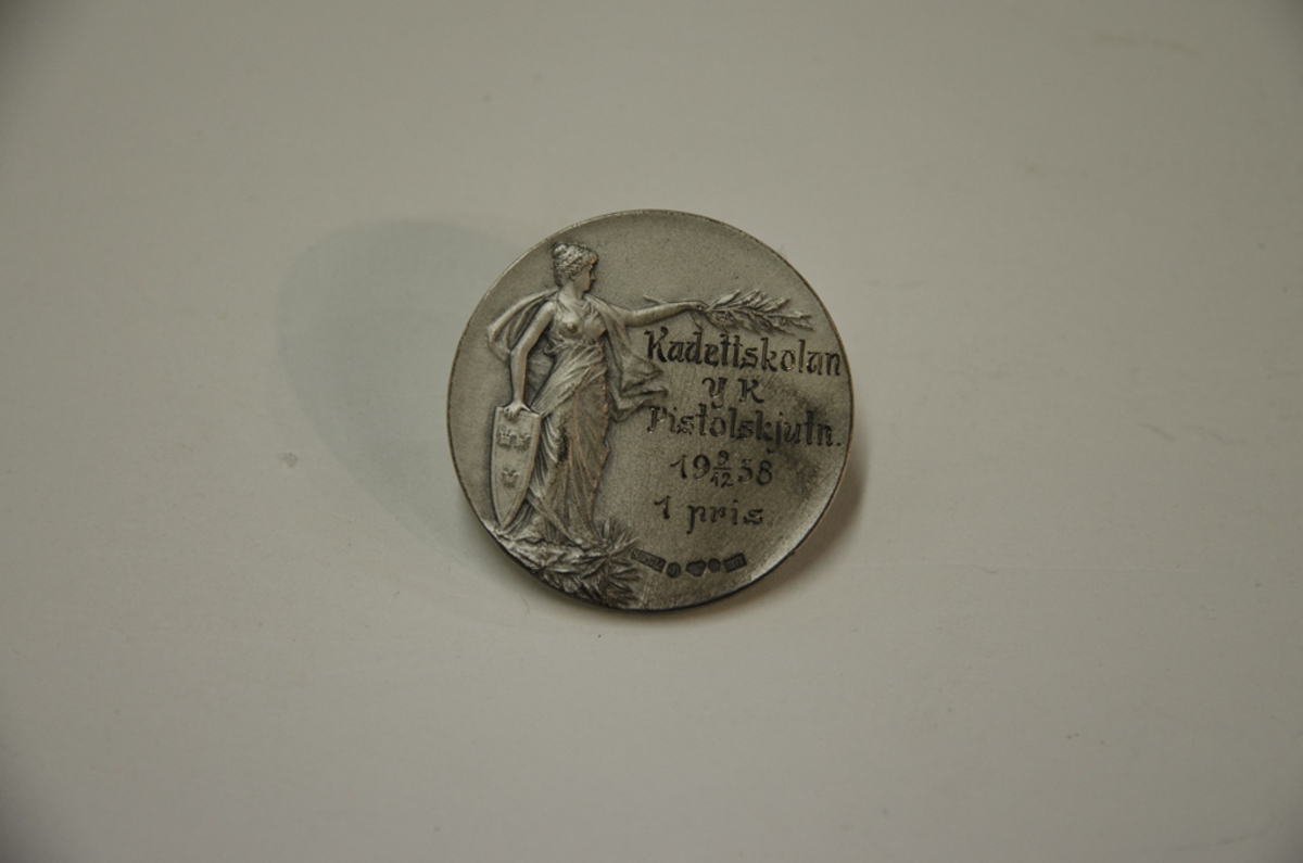 Idrottsmedalj i silver med texten: Kadettskolan YK Pistolskjutn. 9/12 1938, 1 pris. På andra sidan står det: Sveriges Militära Idrottsförbund.