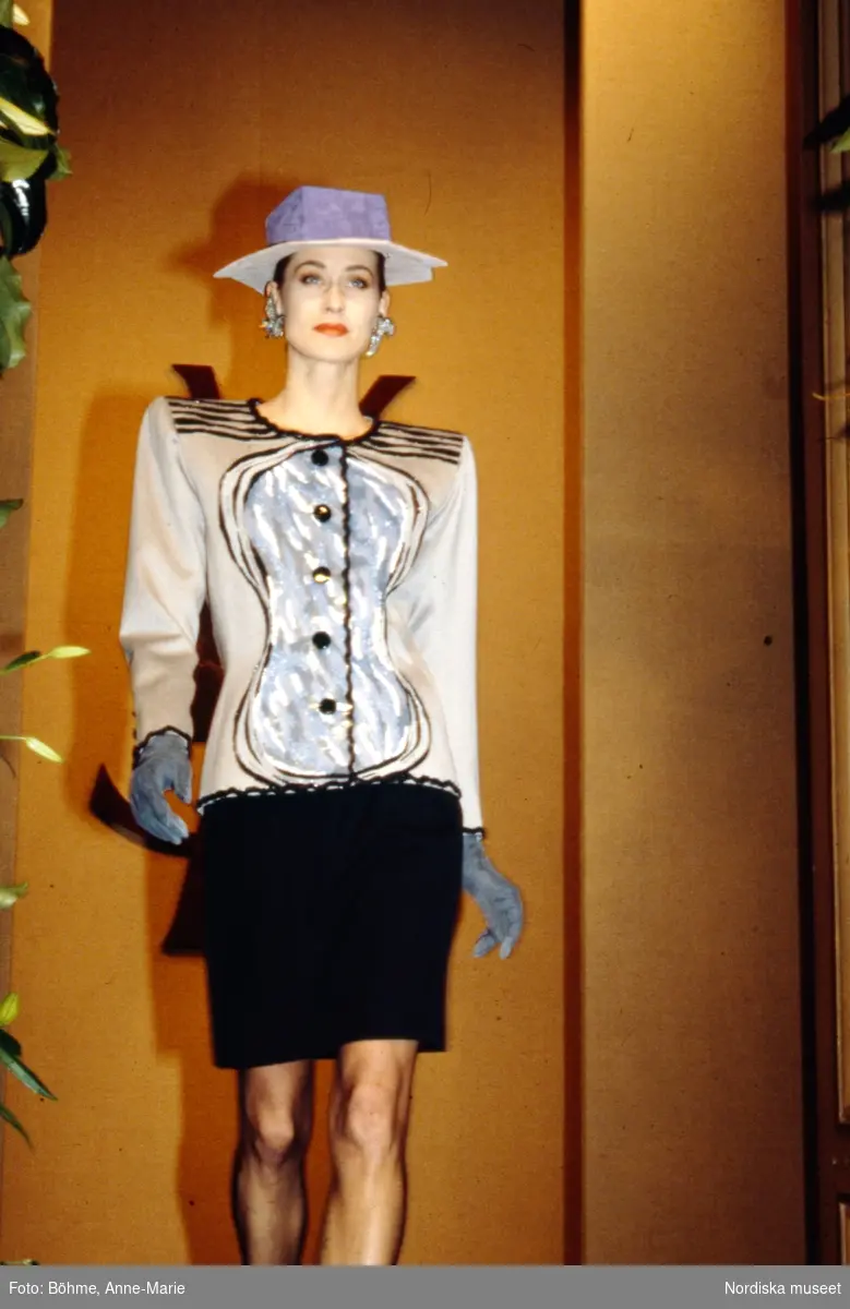 Modevisning. Modell i beige jacka med grått motiv, lila hatt med fyrkantig kulle och svart kjol. Från Yves Saint Laurent.