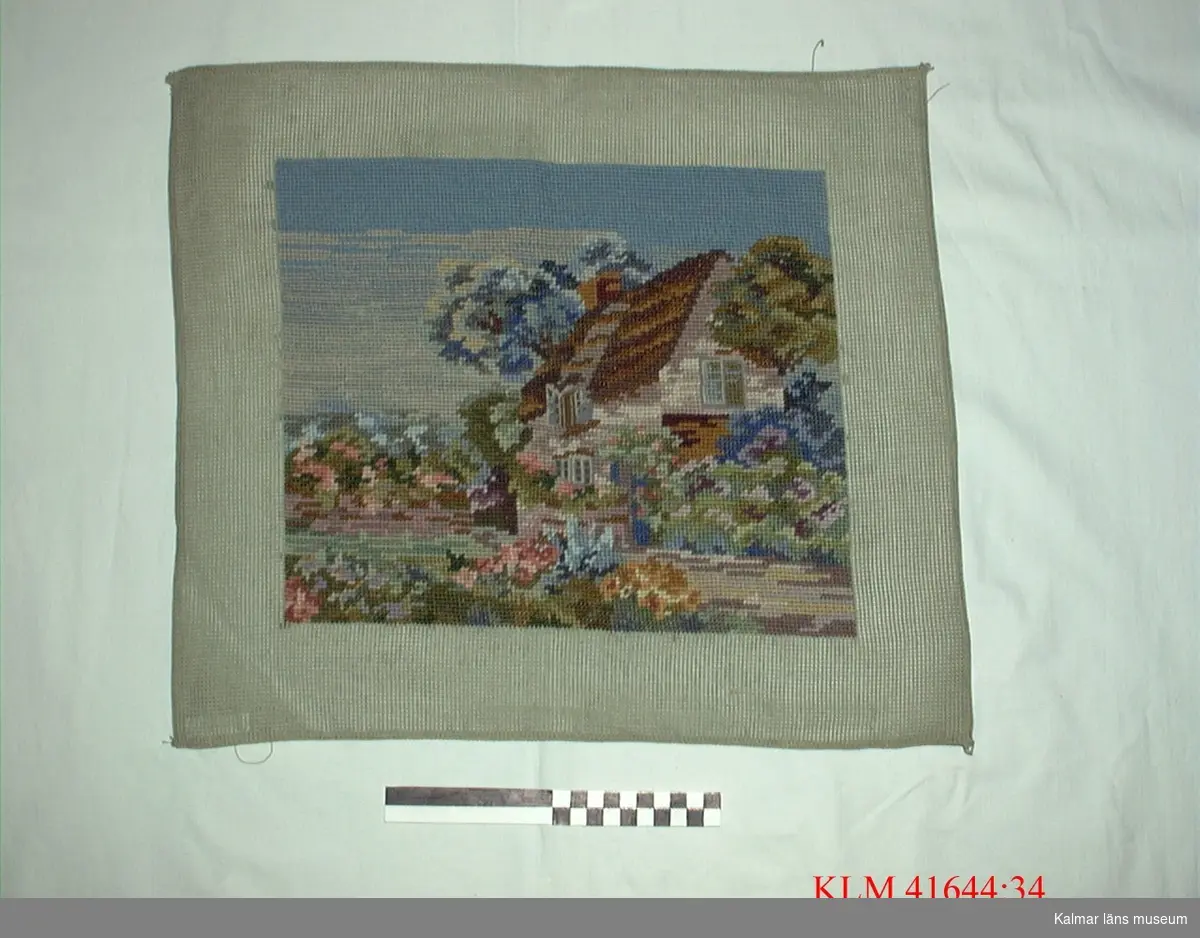 KLM 41644:34 Broderi, textil. Ett nästan kvadratiskt stycke stramalj med fyrkantigt trådbundet broderi, en trädgård med ett hus i färger.
Troligen avsett till tavla.