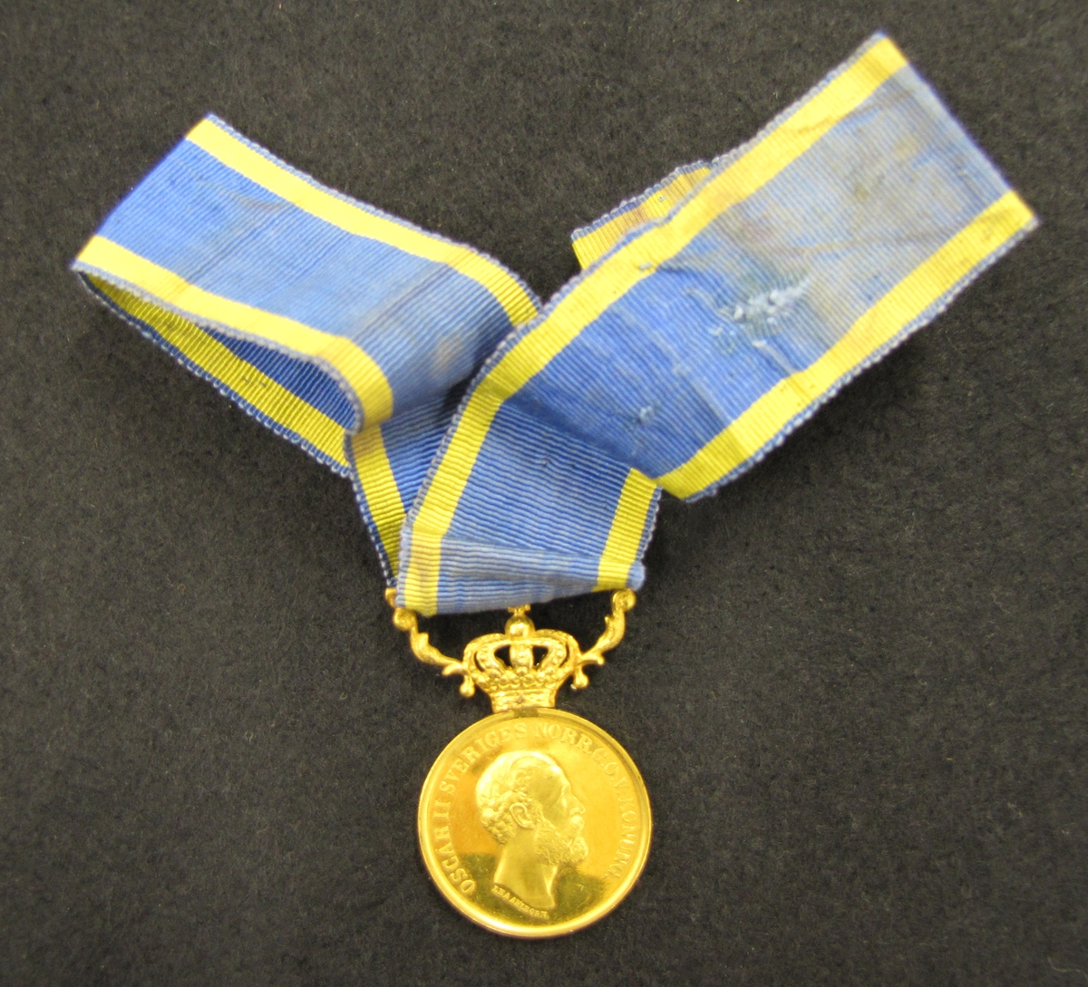 Medalj med blågult band. Medaljen är av 5:e storleken i sitt numera mycket sällsynta originaletui, utdelad 1880 till Lotsen Johan Fredrik Kafvelström (1823-10-03 - 1899-02-07) i Vänersborg. 

Medaljen är av 23 K guld och krona och bärornament av 18 K guld och monterat med sitt originalband vilket Kafvelström av bevarade fotografier bar monterat i en rosett, viket normalt endast brukas av damer.

Medaljen "För berömliga gärningar" instiftad 1803 men kom först till användning 1842 då storlekarna 8 & 5 infördes. Medaljen utdelades i såväl guld som silver. Vilken av dem räddningsmannen tilldelades berodde givetvis på bragdens betydelse och i vilken livsfara även den räddande och den räddade befunnit sig vid olyckan. Den räddandes sociala ställning hade även, märkligt nog, betydelse för vilken av de olika storlekarna som utdelades.

X På framsidan: Oskar II:s bild. På frånsidan: Berömliga gerningar till J F Kafvelström Lots

Bilagor: VM 16978
Tidningsklipp, foto av Lotsen J F Kafvelström, intervju med Arndt Winberg (barnbarn till Kafvelström), bandinspelning.

Släktingarna till Losten J. F Kafvelström. Barnbarn: Arndt, Anna-Greta, Karin och John Winberg, Uddevalla, Barnbarns barn: Frank Larsson, Vänersborg.