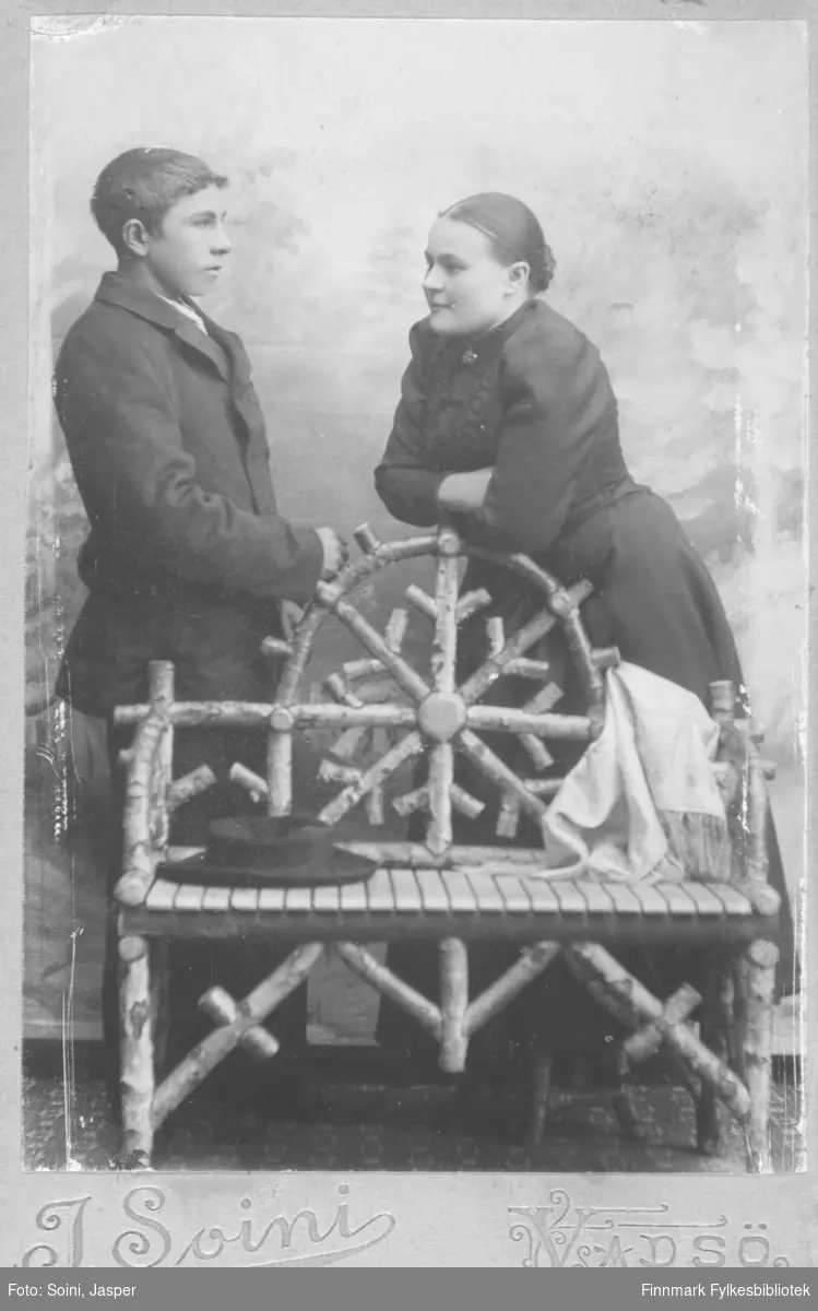 Dobbeltportrett av ukjent par som poserer i fotoatelier med en benk bygget av bjørketre som kulisse. Bakgrunnen er et håndmalt landskap med noe som ligner graner. Kanskje er det et søskenpar? Fotografert av Jasper Soini, Vadsø.