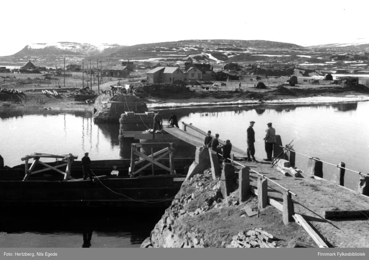 Brubygging i Vestre Jakobselv, 1946. En båt ligger under bruen. En mann er ombord i båten. Fire menn jobber på bruen. Tre gutter står å ser på arbeidet. I bakgrunnen ser man noen hus.