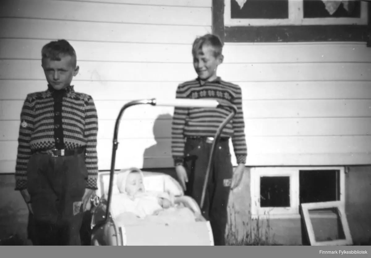 Søsknene Tormod og Svanhild Rushfeldt poserer sammen med onkelen Helge Rystrøm. Svanhild sover i barnevogna med kyse, gutta i stram giv akt kledd i strikkegensere foran et gjenreisningshus med liggende panel. Et kjellervindu står på bakken ved siden av åpningen. Pent vær med sol.