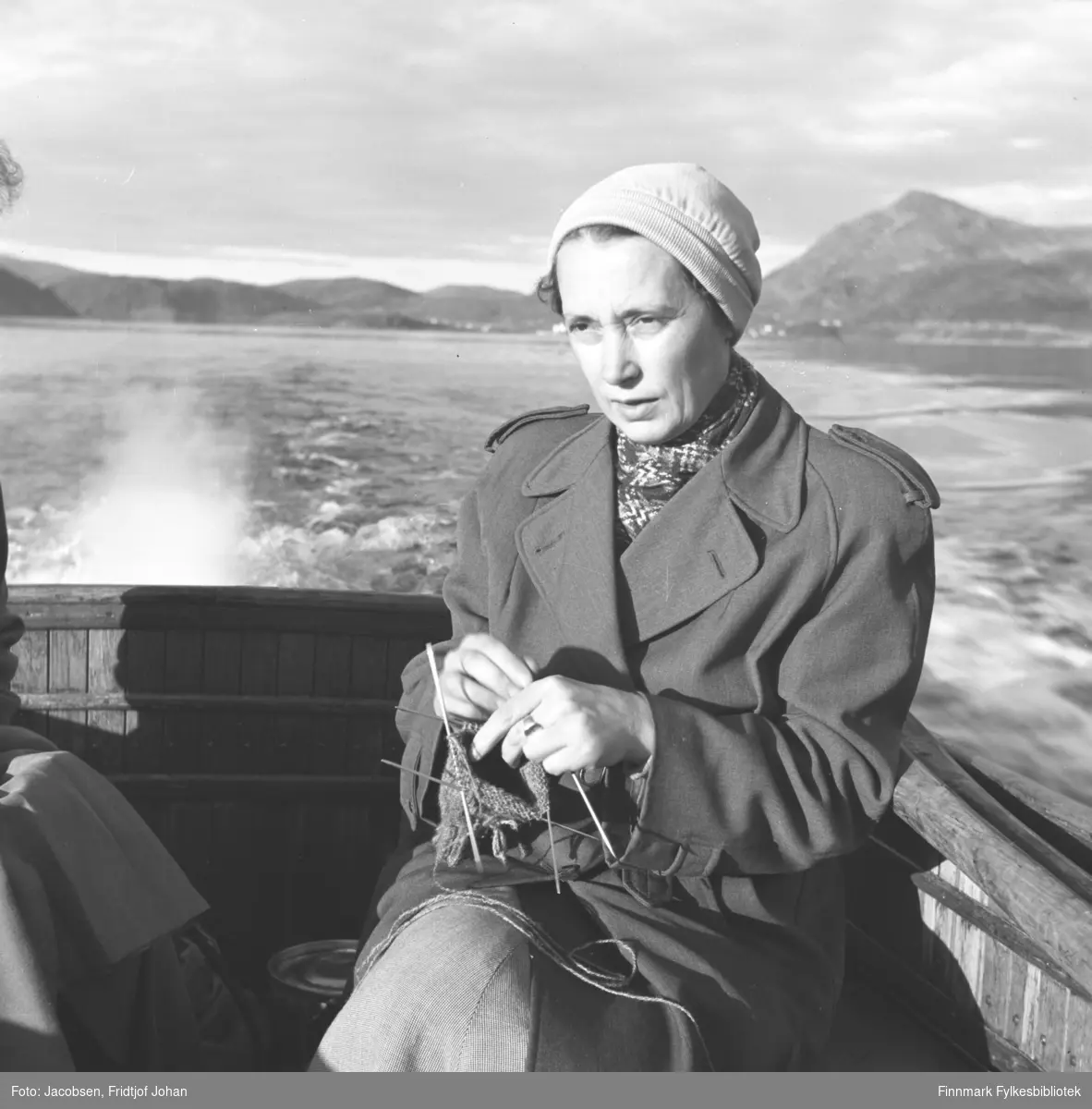 Aase Jacobsen med strikketøyet på båttur i Sørøysundet. Hun sitter på båtdekket iført en ganske mørk kåpe med skjerf i halsen og lys lue på hodet. Kjølvannet og eksosen ses bak henne og bygningene som står i bakgrunnen er Rypefjord.